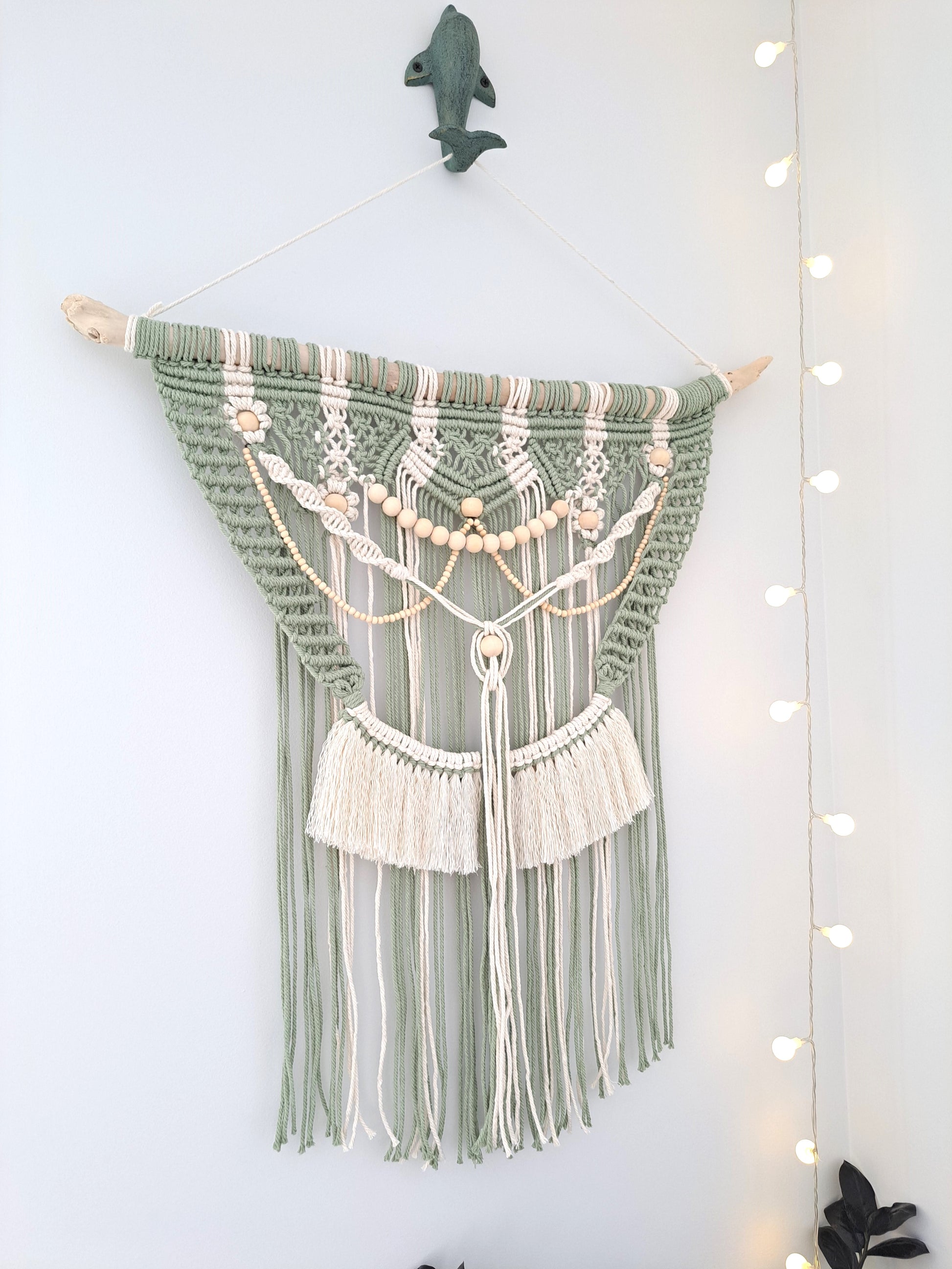 Weathered Landing's Boho Macrame Driftwood Wall Hanging. Handmade with sage green and white cotton macrame, wooden beads, and driftwood, this carefully crafted boho wall hanging is delicate and dainty. Perfect for a nursery or a bedroom nook. With lots of contrast in colour, texture, and pattern, this eclectic macrame wall hanging adds a bohemian feel to the space. Shipping to Canada and US.